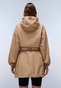 Veste à capuche beige avec une taille cintrée et un ourlet élastique. Dotée de manches bouffantes et d'une ceinture réglable à motifs. Tissu doux et léger.