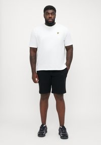 Hvit t-skjorte med korte ermer og et gult logo på venstre bryst, kombinert med svarte shorts og svarte joggesko.