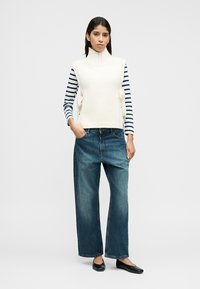 Λευκό πλεκτό τοπ χωρίς μανίκια με ψηλό γιακά, πάνω από μπλε και άσπρη ριγέ μακρυμάνικη μπλούζα. Συνδυασμένο με άνετο σκούρο τζιν και μαύρα φλατ παπούτσια.