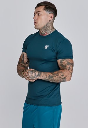 Teal színű, testre simuló sportpóló sima, rugalmas anyagból készült. Kis fehér logó található a mellkason. Hosszú ujjú és kerek nyakú kialakítás.