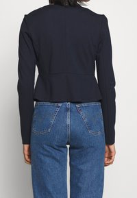 Marineblauwe peplumjas met lange mouwen, gladde textuur en een getailleerde achterkant, gecombineerd met hooggesneden blauwe denimjeans met achterzakken.