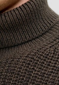 Gros plan sur un pull roulé marron en tricot montrant le col côtelé détaillé et le motif de points texturé sur le tissu.