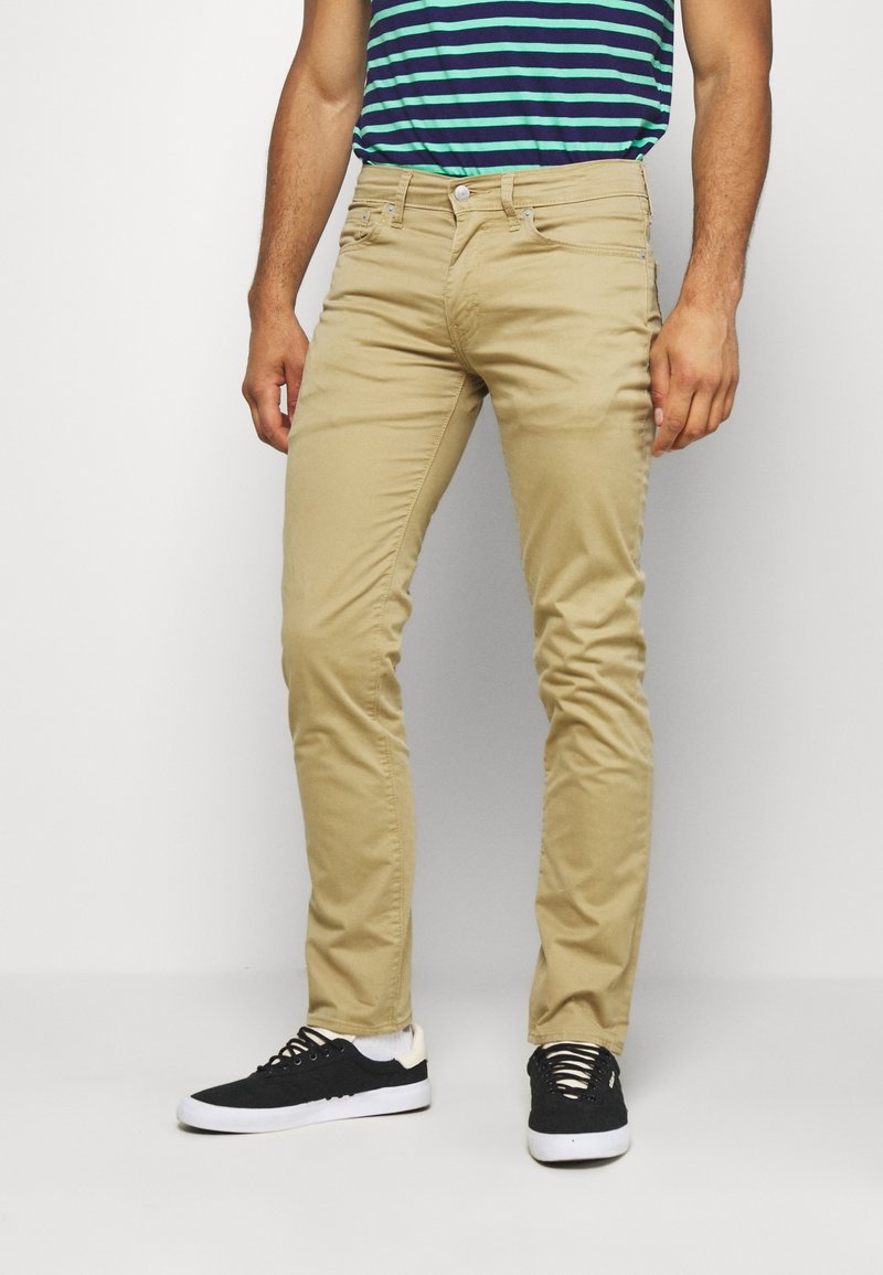 Beige slim-fit chinos met een gladde textuur, voorkeurszakken en een ritsopening. Gecombineerd met zwarte sneakers met witte zolen.