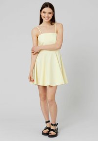 Robe sans manches jaune clair avec un corsage ajusté et une jupe évasée. Présente des fines bretelles et une texture lisse. Portée avec des sandales noires à lanières.