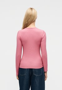 Ξανθός/ή με την πλάτη γυρισμένη, φοράει ροζ εφαρμοστή μπλούζα μακρυμάνικη και άνετο σκούρο μπλε τζιν, με φόντο απλό και ανοιχτόχρωμο.