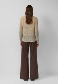 Vrouw met lang bruin haar draagt een beige gebreide trui en een wijde, donkerbruine broek, staand met haar gezicht naar een effen grijze muur.