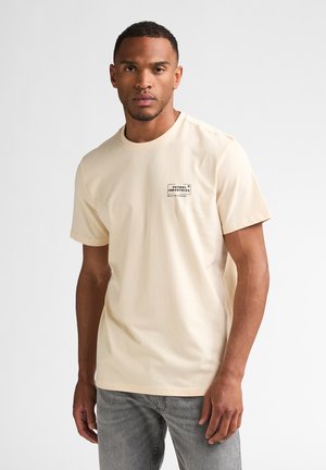 Homme portant un t-shirt beige de Petrol Industries et un jean gris, debout devant un fond clair uni.