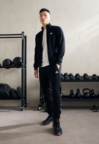 Black athletic jacket and pants made of breathable material, with white logo accents. Standing on a gym floor near weights and equipment.