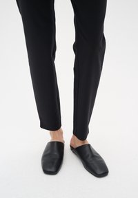 Scarpe slip-on in pelle nera con design a punta quadrata. Indossate con pantaloni neri aderenti, evidenziando una texture liscia e uno stile minimalista.