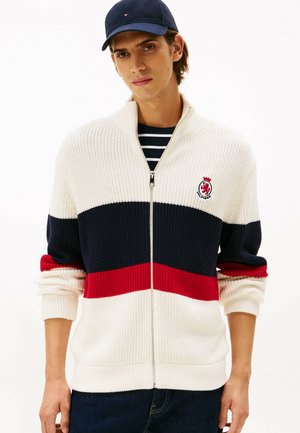 Strick-Zip-up-Cardigan in Creme, Marineblau und roten Streifen; mit geripptem Muster und einem Logo-Patch. Getragen über einem gestreiften Hemd mit Kragen.