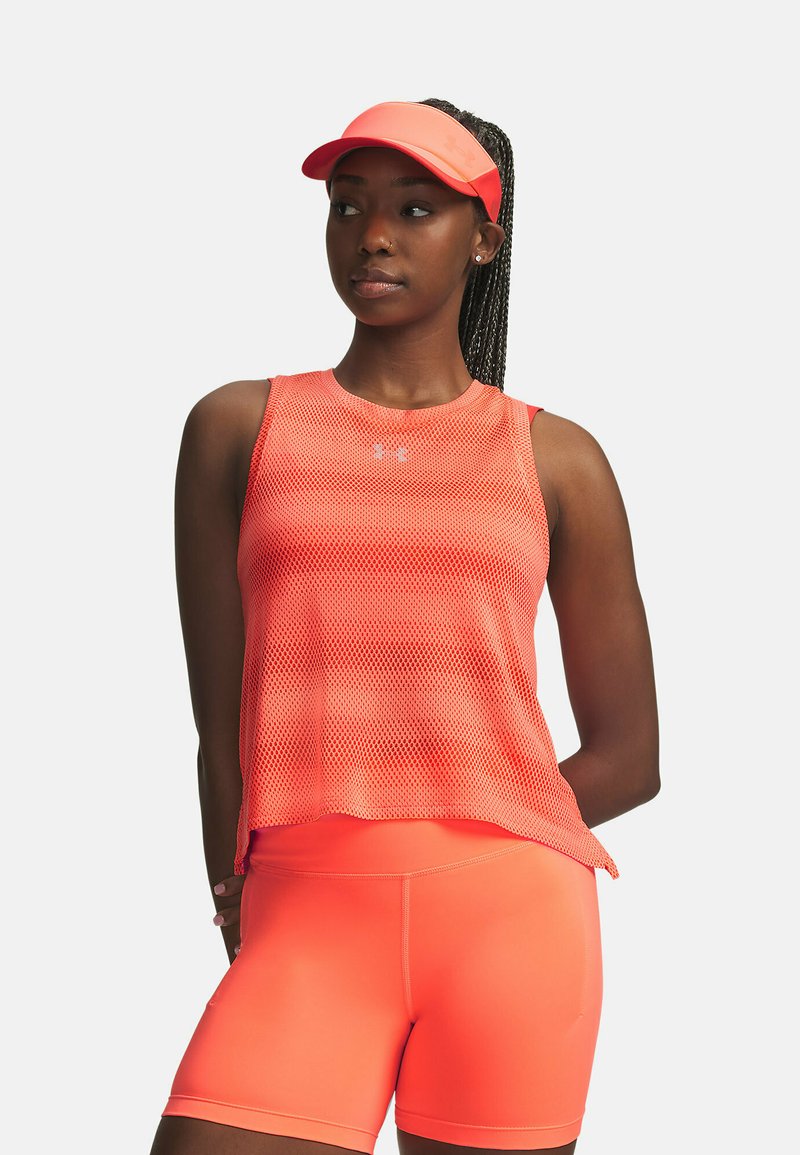 Femme portant un haut sans manches en maille orange vif, un short taille haute assorti et une visière, debout les mains derrière le dos, regardant vers la droite.