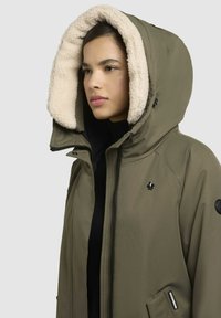 Olijfgroene jas met een grote beige fleece-gevoerde capuchon, voorkant met rits en zijzakken. Bevat een subtiel logo en moderne ontwerp elementen.
