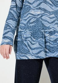 Maglietta a maniche lunghe con un motivo astratto blu e tessuto texturizzato; design a peplo. Abbinata a pantaloni blu scuro con una sottile texture a granellini.