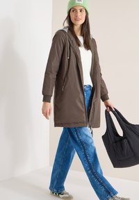 Femme portant un bonnet vert, une veste marron, un pull blanc, un jean bleu, tenant un sac noir matelassé, marchant devant un fond beige et blanc.