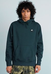 Jovem com cabelo encaracolado, vestindo um hoodie da Nike Air verde-escuro e calças de camuflagem, de pé contra um fundo claro e simples.
