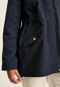 Tumši zils džemperis, kas izgatavots no viegla auduma, ar saitēm viduklī, priekšējo kabatu ar zelta pogu un gludu tekstūru.