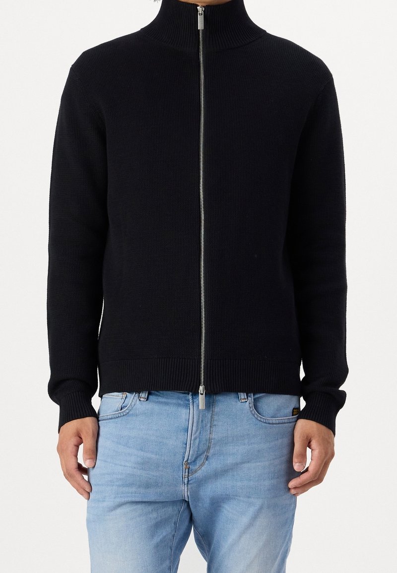 Svart zip-up-tröja med hög krage, ribbad struktur och åtsittande manschetter. Matchas med ljusblå jeans som har en diskret logotyp på fickan.