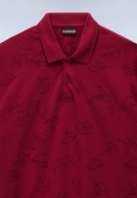 Mörkröd polo skjorta med ett strukturerat bladmönster, kort krage och knäppning med tre knappar. Har en broderad logotyp på vänster bröst.