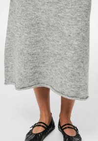 Vestido midi de punto gris con una textura suave, que presenta un dobladillo ligeramente ondulado. Combinado con zapatos planos negros con dobles tiras y un acento decorativo.