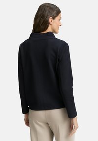 Woman with brown hair wearing a textured black long-sleeve jacket and beige pants, facing away, against a plain white background.