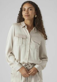 Chemise beige à boutons en tissu doux et lisse, dotée de deux poches poitrine, de manches longues et d'une coupe décontractée. Détail du bas avec cordon de serrage visible.