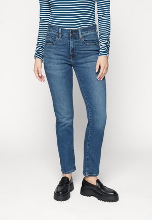 Persoon draagt een blauw-wit gestreept shirt met lange mouwen, blauwe jeans en zwarte loafers, staand tegen een effen witte achtergrond.