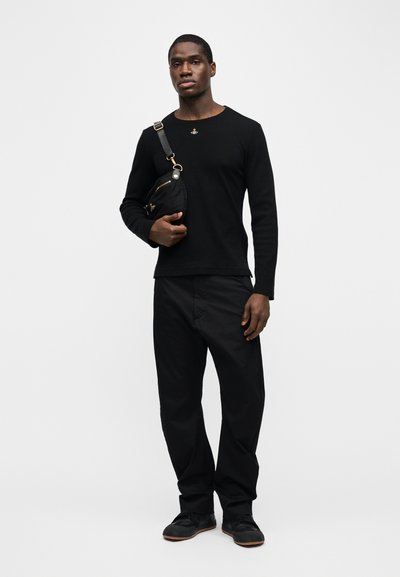 Άντρας που στέκεται φορώντας μαύρο μακρυμάνικο πουκάμισο, μαύρο παντελόνι, μαύρα παπούτσια και κρατώντας μια μαύρη τσάντα χιαστί μπροστά σε απλό λευκό φόντο.