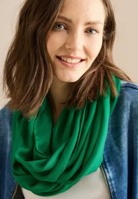 Écharpe verte, légère, en tissu doux, drapée en couches. Portée avec une veste en jean, présentant une texture lisse et une couleur vive.