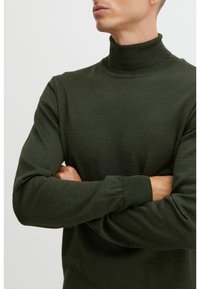 Hombre que lleva un suéter de cuello alto verde oscuro con los brazos cruzados, visto de cuello a cintura sobre un fondo liso.