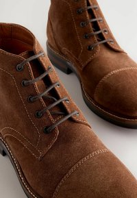 Braune Wildleder-Ankle-Boots mit runder Spitze, runden Schnürsenkeln, kontrastierenden Nähten und einer strukturierten Oberfläche. Gummisohle mit einem leichten Absatz.