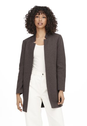 Cappotto lungo marrone, aperto davanti, realizzato in tessuto spesso. Presenta un design semplice, scollo rotondo e maniche lunghe. Indossato sopra un top bianco.
