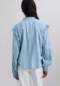 Blusa azul clara con tejido texturizado, hombros con volantes, mangas largas con puños fruncidos y un dobladillo ligeramente curvado.