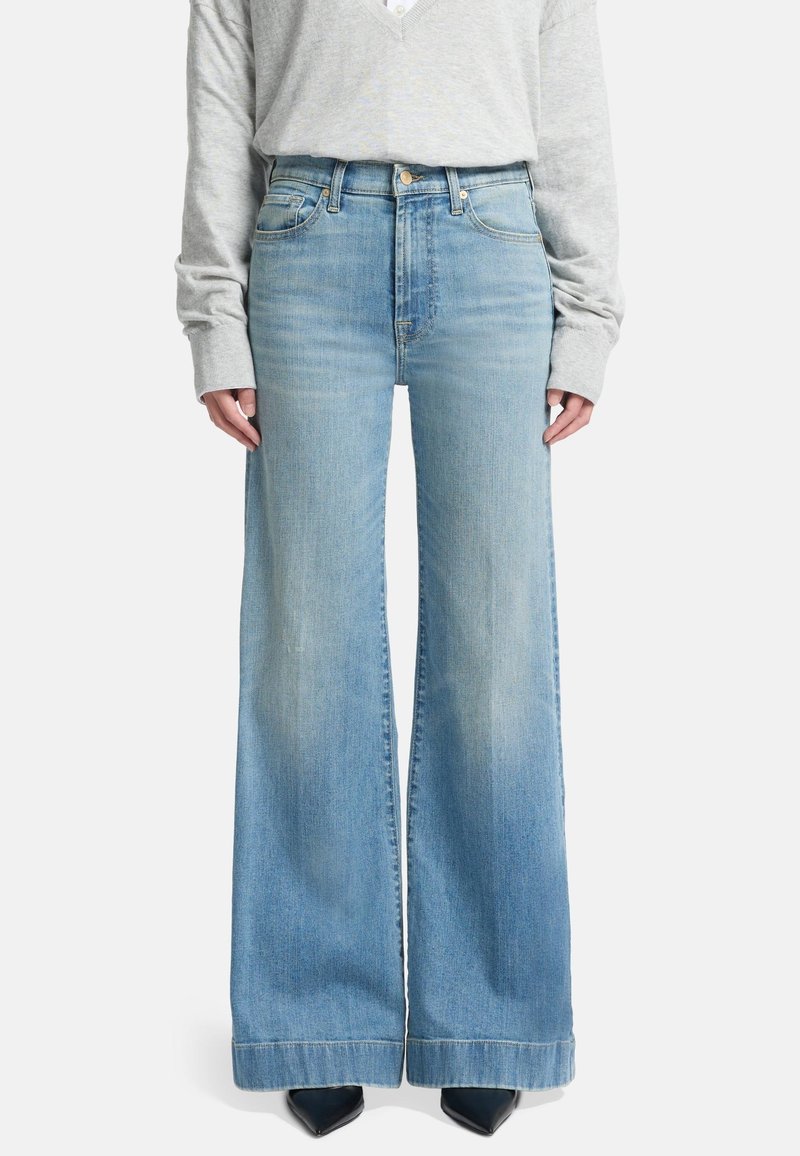 Wide-leg jeans in light blue denim, featuring a high waistband and a faded finish, paired with a gray long-sleeve top and black heels.