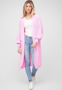 Cardigan lungo lavorato a maglia di un brillante rosa, con maniche a palloncino, apertura frontale e tasche anteriori, abbinato a jeans skinny azzurri e scarpe da ginnastica bianche.