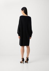 Black oversized knit dress with long sleeves, featuring a wide neckline and smooth texture, paired with black high-heeled shoes.