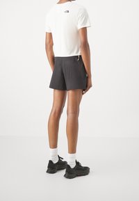 Vit kortärmad skjorta med logotyp på ryggen, kombinerad med mörka shorts och svarta sportskor. Enkel, funktionell design.