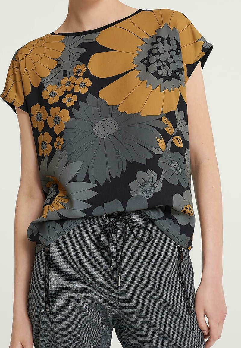Blusa de manga corta con estampados amarillos y grises sobre un fondo negro, combinada con pantalones grises texturizados que presentan detalles con cremallera.