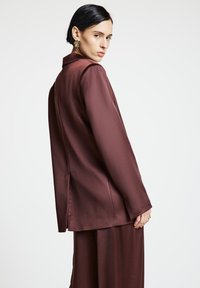 Frau mit kurzen schwarzen Haaren, gekleidet in einen passenden dunkel mauvefarbenen Satinblazer und Hose, seitlich stehend, mit goldenen, blumenförmigen Ohrringen.