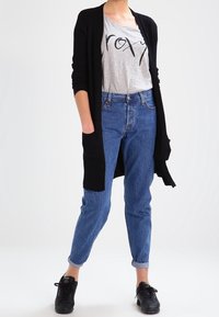 Schwarze gerippte Strickjacke, graues T-Shirt mit schwarzem Grafikdruck, hoch taillierte blaue Jeans und schwarze Schnürschuhe.