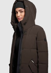 Brun oppustet jakke med hætte, der har en lynlås lukning, trykknapper og forlomme. Teksturstof med en mat finish.