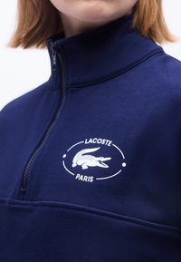 Gros plan d'une personne portant un haut à demi-zip bleu marine avec un logo blanc Lacoste Paris représentant un crocodile sur la poitrine.
