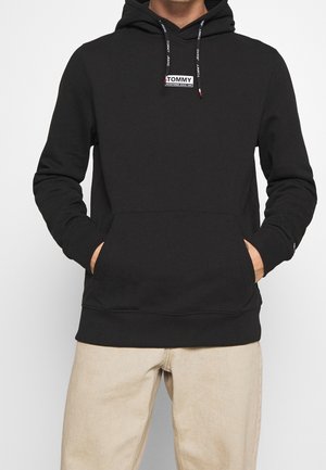 Person in einem schwarzen Kapuzenpullover mit "Tommy"-Logo auf der Brust, mit Schnürsenkeln und Text, sowie beigen Hosen, Hände in der vorderen Tasche.