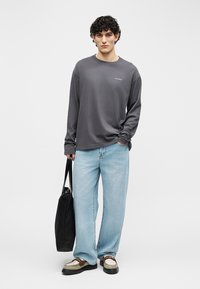 Jonge man met krullend haar, gekleed in een donkergrijs shirt met lange mouwen, lichtblauwe brede jeans, bruine en witte schoenen, en draagt een zwarte totebag.