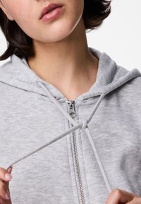 Sivi dukserica s patentnim zatvaračem i kapuljačom na vezanje. Mekana tekstura, rebrasti ovratnik i metalni detalj patenta. Blizu pogled na izrez i kapuljaču.