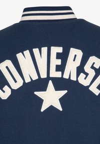Giacca in stile universitario blu navy con lettere ricamate bianche che formano la parola "CONVERSE" e una stella, dotata di un colletto a righe e una texture morbida.