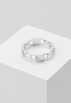Bague en maillons de chaîne argentée avec sections triangulaires serties de petites pierres transparentes, posée sur un socle cubique blanc sur fond gris.