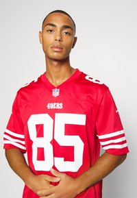 Piros NFL mez fehér díszítéssel, 85-ös számmal és logóval. Rövid ujjak csíkozott részletekkel a mandzsetták közelében. Sima anyagtextúra.