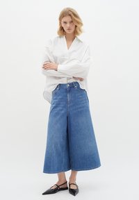 White oversized blouse with long sleeves, paired with wide-leg blue denim culottes. Model wears black pointed-toe flats.