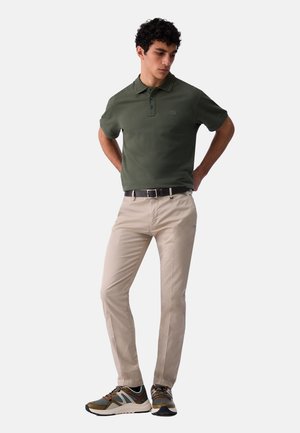 Uomo che indossa una polo verde scuro, pantaloni beige con cintura nera e scarpe da ginnastica multicolori, in piedi con le mani dietro la schiena.