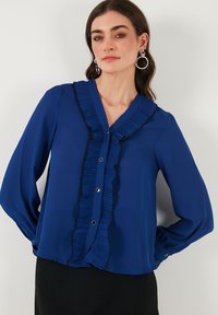 Blue blouse made of smooth fabric, featuring a v-neck with pleated trim, black accents, and a button-down front. Long sleeves complete the design.